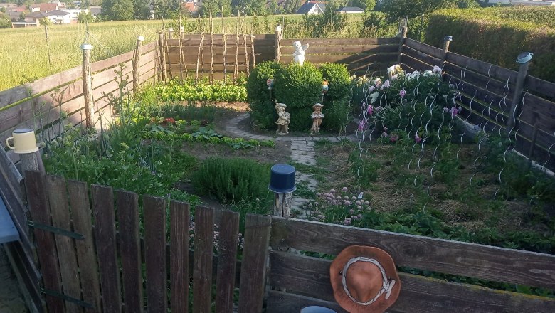 img_20210614_183455_ausschn_sonnenkraeuterhof-schmidt, © Sonnenkräuterhof Schmidt Ein gepflegter Garten mit Gemüsebeeten, Blumen und Dekorationen, umgeben von einem Holzzaun. Im Hintergrund ist eine Kirche und eine Stadt zu sehen.