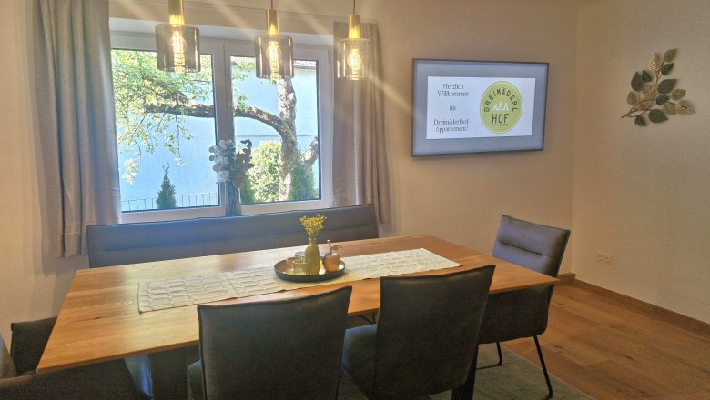 Dreimäderlhof, © Helga und Thomas Kraushofer Esszimmer mit Holztisch, Stühlen und Fenster mit Blick auf einen Baum. An der Wand hängt ein Bildschirm mit Begrüßungstext.