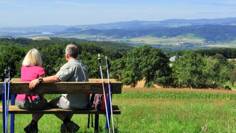 Durch die hügelige Landschaft wandern, © ARGE Dunkelsteinerwald Ein Paar sitzt auf einer Bank mit Wanderstöcken und blickt auf eine hügelige Landschaft.