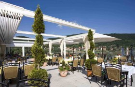 Residenz Wachau - Terrasse, © Hotel Donauterrasse Terrasse der Residenz Wachau mit Tischen und Stühlen, umgeben von Pflanzen, mit Blick auf einen Fluss und grüne Hügel im Hintergrund.