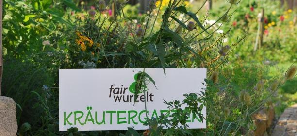 Kräutergarten fairwurzelt, © fairwurzelt Ein Schild mit der Aufschrift 'fairwurzelt Kräutergarten' steht inmitten von grünen Pflanzen und Blumen.
