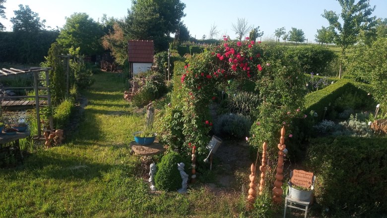 img_20210614_183801_klein_sonnenkraeuterhof-schmidt, © Sonnenkräuterhof Schmidt Ein gepflegter Garten mit Rosenbogen, grünen Hecken und dekorativen Elementen bei Sonnenschein.