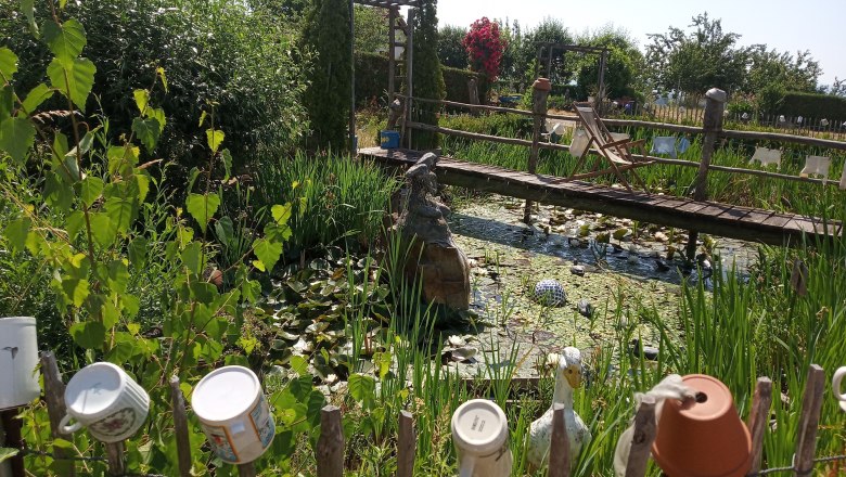 img_20210624_104052_klein_sonnenkraeuterhof-schmidt, © Sonnenkräuterhof Schmidt Ein Garten mit Teich, Brücke und Tassen auf einem Zaun.