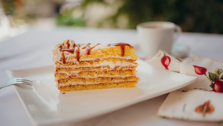Hagebuttentorte, © Niederösterreich Werbung/Daniela Führer Ein Stück Schichttorte mit Sahne und Karamell auf einem weißen Teller, daneben eine Tasse Kaffee und eine Serviette mit Fruchtmuster.