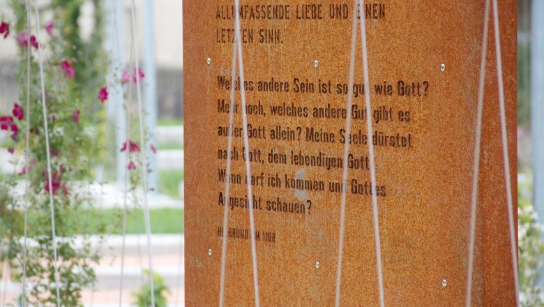 Texte regen zum Nachdenken an, © Anna Lun Nahaufnahme einer rostigen Metalltafel mit eingraviertem Text, umgeben von Pflanzen.