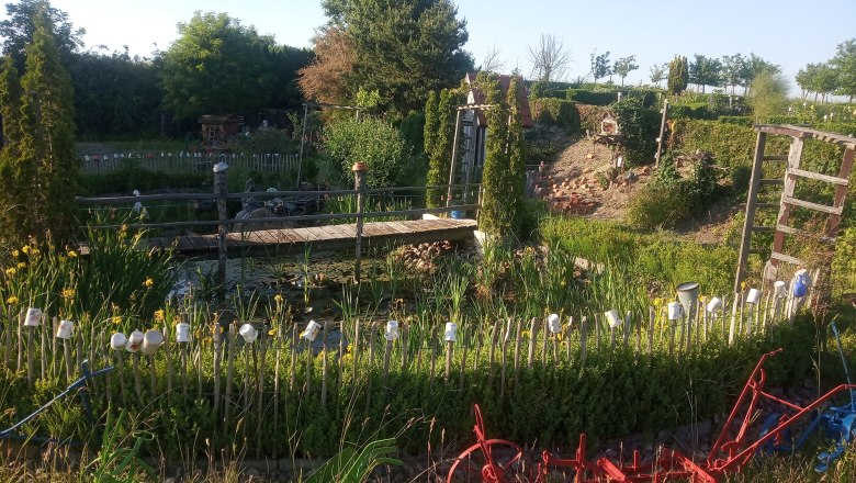 img_20210614_185039_klein_sonnenkraeuterhof-schmidt, © Sonnenkräuterhof Schmidt Ein Garten mit Teich, Brücke und dekorativen Tassen auf einem Zaun. Im Vordergrund rote landwirtschaftliche Geräte.