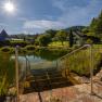 Hotel ResidenzWachau, © Niederösterreich Werbung / Maximilian Pawlikowsky Treppen führen in einen Teich mit klarem Wasser, umgeben von grüner Landschaft und einem Haus im Hintergrund, unter einem strahlend blauen Himmel mit Sonne.