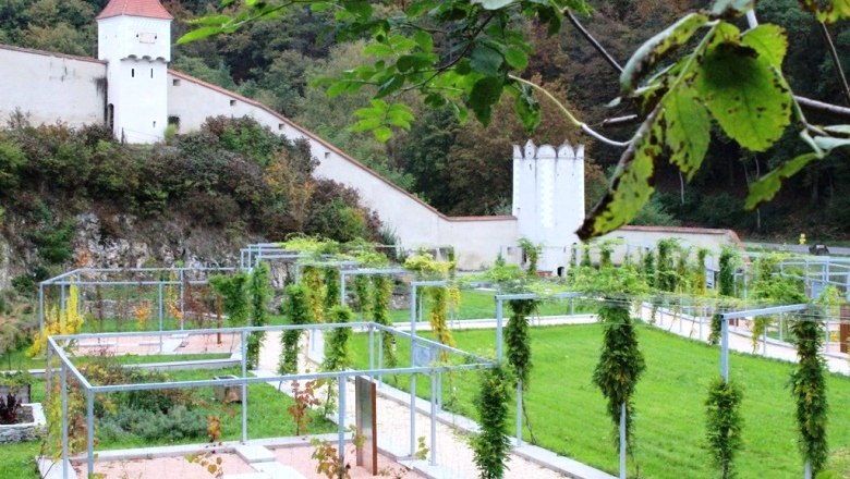 Gartenanlage mit Metallstrukturen und Burgmauer im Hintergrund.