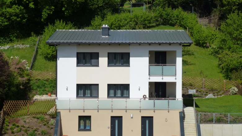 Zweist&ouml;ckiges modernes Haus mit Balkon, umgeben von gr&uuml;ner Landschaft.