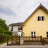 Ein gelbes Haus mit Spitzdach und Holzzaun in einer ländlichen Umgebung.