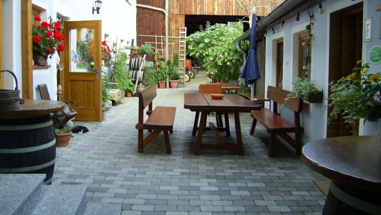Innenhof eines Bauernhofs mit Holztischen und Bänken, umgeben von Pflanzen und Blumen.