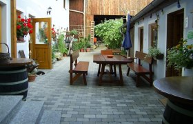 Innenhof eines Bauernhofs mit Holztischen und Bänken, umgeben von Pflanzen und Blumen.